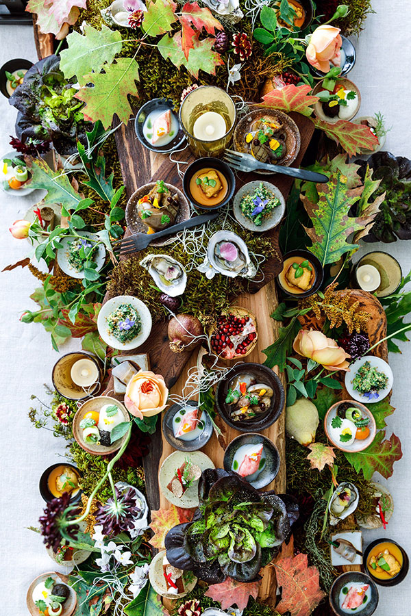 A large table setting with an array of colorful dishes of food surrounded by decorative leaves, flowers, and moss.